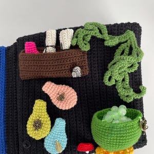 May include: A black crocheted textile with various colorful crocheted appliques. Features include a brown basket with pink and white items, green vines, pear shapes, a green bowl with beads, and a red mushroom.
