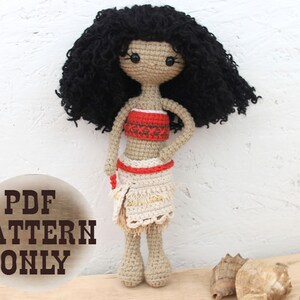 Könnte beinhalten: Häkelanleitung für eine Puppe mit schwarzen Locken, einem rot-weißen Oberteil und einem beigefarbenen Rock. Die Puppe steht auf einer Holzoberfläche mit Muscheln.
