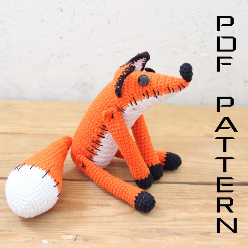 May include: Crocheted orange fox plush toy with black accents and white tail. The toy is sitting on a wooden surface. PDF PATTERN.