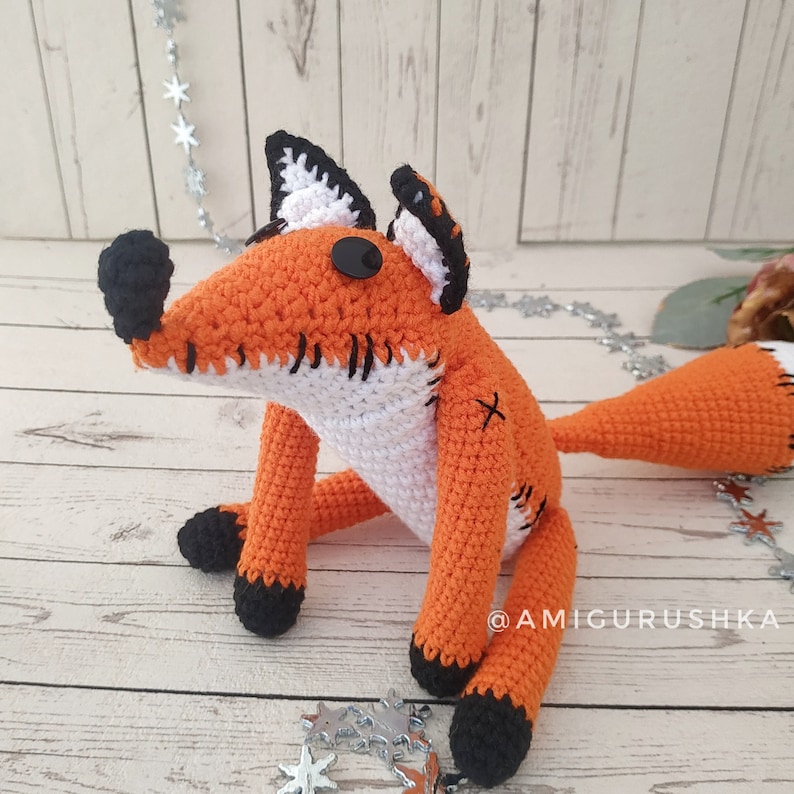 May include: A crocheted fox plush toy with orange and white fur, black eyes, and a black nose. The toy has stitched details on its body and a black stitched X on its back. The toy is sitting on a wooden surface with silver stars scattered around it. The text "@AMIGURUSHKA" is visible in the lower right corner of the image.