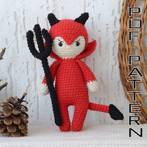 Puede incluir: Un disfraz de diablo de crochet rojo con cuernos y cola negros. El diablo sostiene un tridente negro.