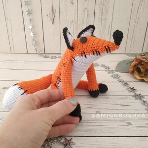 May include: A crocheted orange and white fox plush toy with black eyes and nose. The fox is sitting on a white wooden surface with a silver garland of stars in the background.