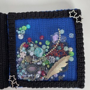 May include: A handmade journal with a black crocheted border and a blue mesh center filled with beads, charms, and a gold feather. Silver star accents adorn the corners, creating a unique, artistic design.