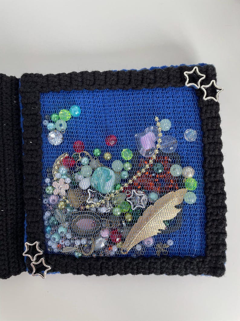 May include: A handmade journal with a black crocheted border and a blue mesh center filled with beads, charms, and a gold feather. Silver star accents adorn the corners, creating a unique, artistic design.
