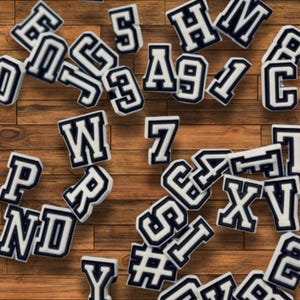 May include: A selection of white and navy blue varsity-style letter and number patches scattered on a wooden surface. The patches include letters like "W", "P", "S", and numbers like "7" and "9".