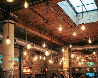 May include: A crowded bar with exposed brick walls and a wooden ceiling. The bar is lit by many hanging Edison light bulbs. There are people of all ages and races in the bar, enjoying drinks and conversation.