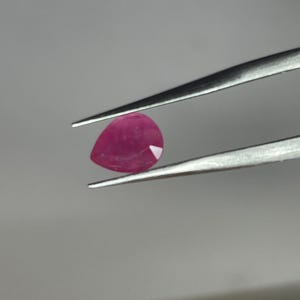 May include: A pear-shaped, deep red ruby gemstone held by silver tweezers. The gemstone has a polished surface and a slight facet on one side, reflecting light. The background is a neutral gray.