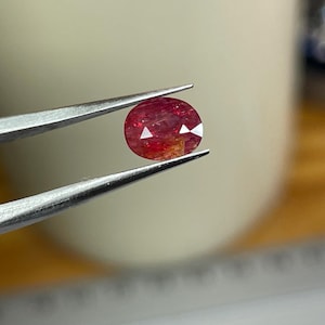Könnte beinhalten: Ein oval geschliffener Rubin-Edelstein, der von einer Pinzette gehalten wird. Der Rubin hat eine tiefrote Farbe mit helleren orangefarbenen und weißen Einschlüssen. Die Facetten reflektieren das Licht und erzeugen einen funkelnden Effekt.