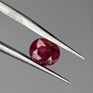 Könnte beinhalten: Ein facettierter, ovalförmiger Rubin, gehalten von einer silbernen Pinzette. Der tiefrote Edelstein weist sichtbare Einschlüsse auf. Der Hintergrund ist grau, wodurch die Brillanz des Rubins hervorgehoben wird.