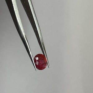 Könnte beinhalten: Ein facettierter, ovalförmiger Rubin-Edelstein, der von einer silbernen Pinzette gehalten wird. Der Rubin zeigt eine tiefrote Farbe mit inneren Mustern. Die Pinzette ist vor einem schlichten, hellgrauen Hintergrund positioniert.