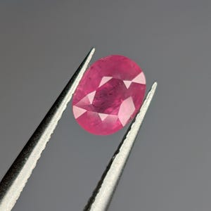 Könnte beinhalten: Ein leuchtend rosa ovaler Edelstein, der von einer Pinzette gehalten wird. Der Edelstein hat einen facettierten Schliff und eine glänzende Oberfläche.