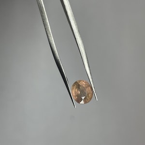 Könnte beinhalten: Ein facettierter, ovaler Edelstein in Pfirsich- und Brauntönen wird von einer silbernen Pinzette vor einem grauen Hintergrund gehalten. Der Schliff des Edelsteins zeigt innere Facetten, die seine Brillanz und Farbe verstärken.