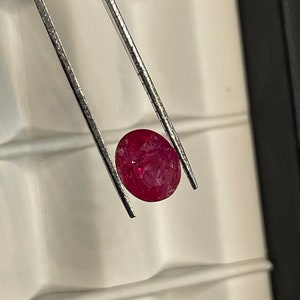 May include: A vibrant red oval gemstone held by silver tweezers. The gemstone has a slightly rough texture and is set against a white background.