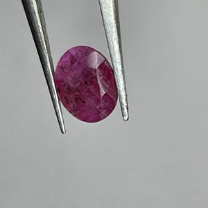 Peut inclure: Une pierre précieuse rubis ovale et facettée, tenue par une pince argentée. Le rubis est d'un rose profond et saturé, avec des inclusions internes et une surface polie. La pierre est présentée sur un fond gris clair uni.