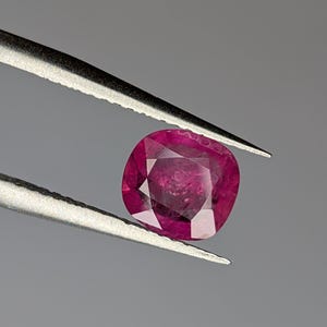 Puede incluir: Una gema rosa vibrante con un corte de cojín, sostenida por pinzas sobre un fondo gris.