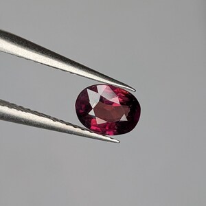 Könnte beinhalten: Ein facettierter, ovaler Edelstein, der von einer Pinzette gehalten wird. Der Edelstein hat eine tiefe, satte rote Farbe mit inneren Facetten, die das Licht reflektieren. Die Pinzette ist silberfarben und der Hintergrund ist ein neutrales Grau.