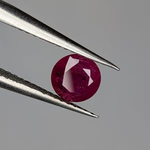 May include: A faceted, round ruby gemstone held by silver tweezers. The ruby is a deep, rich red color with visible internal facets. The tweezers are positioned against a neutral gray background.