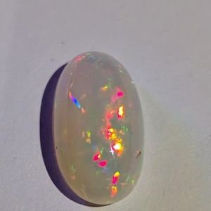 May include: A white oval opal gemstone with iridescent flashes of colour, including pink, green, and blue.