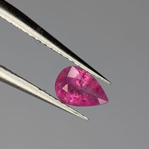 May include: A pear-shaped pink gemstone held by tweezers. The gemstone is faceted and has a brilliant shine.