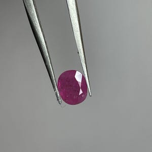 Op de afbeelding: Een gefacetteerde, ovaalvormige robijn edelsteen, vastgehouden door een zilveren pincet. De robijn heeft een diepe, verzadigde roze-rode kleur, met zichtbare interne insluitsels. De pincet bevindt zich tegen een effen, lichtgrijze achtergrond.