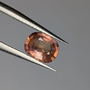 Könnte beinhalten: Ein ovaler Edelstein in hellrosa, der mit einer Pinzette gehalten wird. Der Edelstein hat einen leichten Braunton.