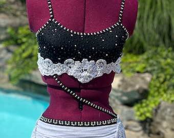 May include: A black and white dance costume on a maroon mannequin. The top is black with silver embellishments and straps. The waist is accented with a white, embellished belt. A white skirt and black bottoms complete the outfit. The background is a pool and greenery.