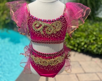 May include: A vibrant fuchsia dance costume with gold embellishments and sparkling accents. The two-piece outfit features a bedazzled crop top and matching bottoms, with sheer, ruffled sleeves. The costume is displayed on a mannequin.
