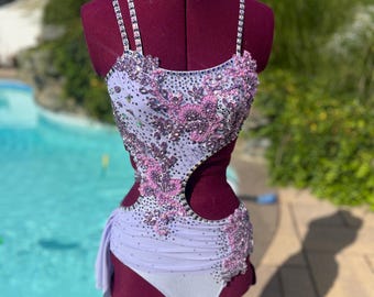 May include: A lavender and white leotard with intricate sequin and rhinestone embellishments. The leotard features a cutout design on the side and a flowing fabric detail. The straps are also adorned with rhinestones. The garment is displayed on a maroon dress form.