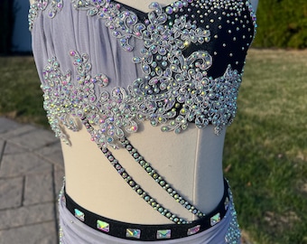 May include: A dance costume with a black and purple bodice and skirt. The bodice is decorated with iridescent rhinestones and silver trim. The skirt is made of a sheer fabric and has a black waistband.