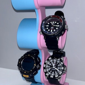 May include: A three-tiered watch stand with a pink and blue color scheme. The stand has two slots on the top tier, one slot on the middle tier, and a base with a pink and blue color scheme. Three watches are displayed on the stand.