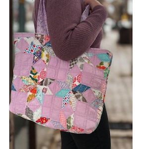 May include: A quilted tote bag featuring a patchwork star pattern in shades of lavender, pink, blue, and white. The bag has wide shoulder straps made from a light purple fabric with a subtle tonal floral print and a soft, stitched texture.