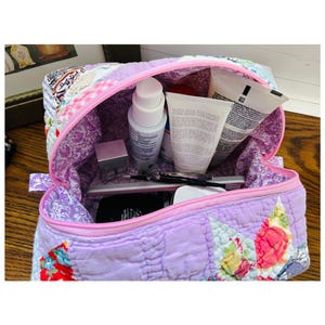 May include: A lavender quilted cosmetic bag with a pink zip, open to show beauty products. The bag has a patchwork design with floral patterns and a quilted texture, with various beauty products inside.