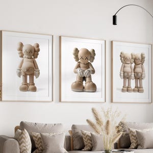 May include: Three framed prints showcasing beige cartoon figures with X-shaped eyes and gloved hands. The figures are in various poses, set against a white backdrop. The frames are light brown, and the artwork is displayed above a grey sofa.