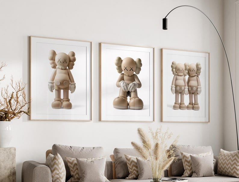 May include: Three framed prints showcasing beige cartoon figures with X-shaped eyes and gloved hands. The figures are in various poses, set against a white backdrop. The frames are light brown, and the artwork is displayed above a grey sofa.