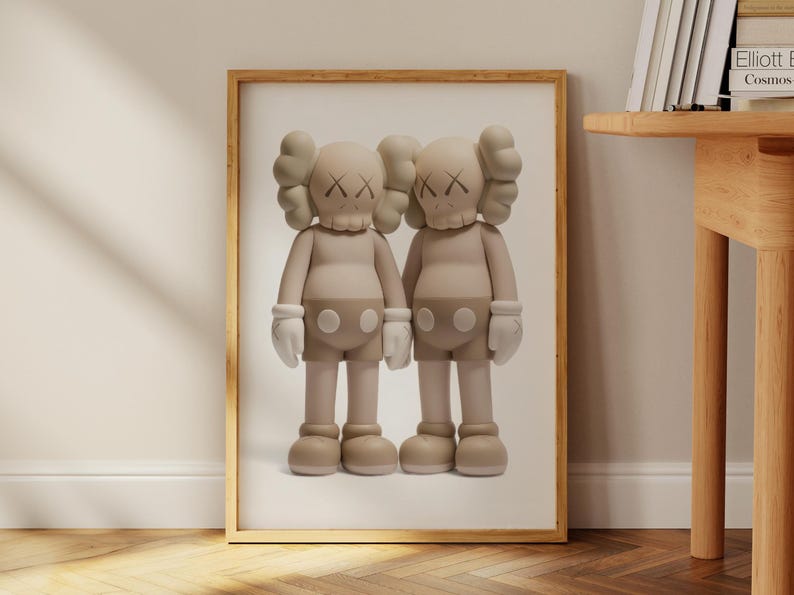 May include: Framed artwork showing two beige cartoon figures with X-shaped eyes. The figures are side by side, wearing white gloves and shorts. The artwork is in a wooden frame, leaning against a wall. The frame is approximately 60 cm in height.