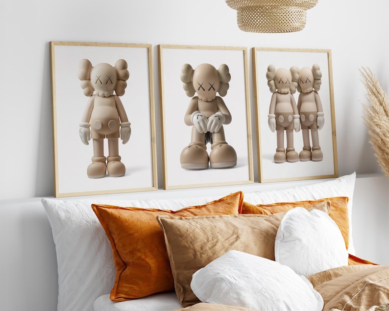 May include: Three framed prints showcasing beige cartoon figures with X-shaped eyes and hands. The figures are in different poses, set against a white backdrop. The frames are light brown, and the prints are displayed above orange and brown cushions.