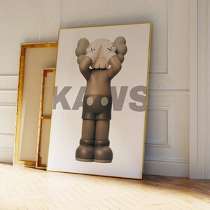 May include: A framed print featuring a brown and gray cartoon character with its hands covering its eyes. The character is standing on a white background with the word "KAWS" printed in gray letters.