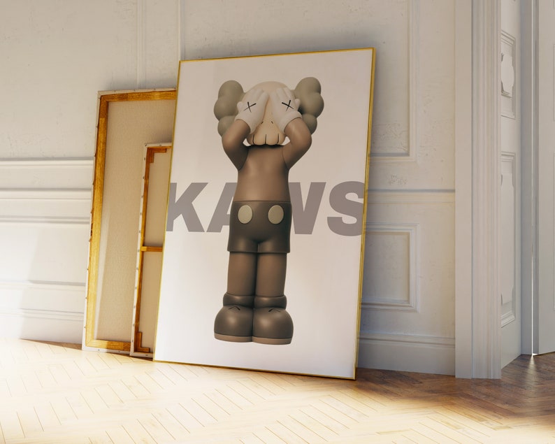 May include: A framed print featuring a brown and gray cartoon character with its hands covering its eyes. The character is standing on a white background with the word "KAWS" printed in gray letters.