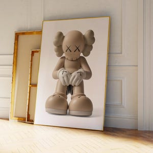 May include: A framed print of a beige cartoon figure with crossed-out eyes and gloved hands. The figure is in a seated position. The print is leaning against a wall next to two other framed artworks. The artwork has a gold frame.