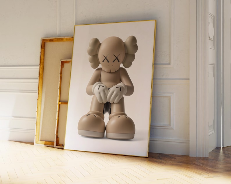 May include: A framed print of a beige cartoon figure with crossed-out eyes and gloved hands. The figure is in a seated position. The print is leaning against a wall next to two other framed artworks. The artwork has a gold frame.