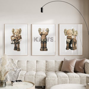 May include: Three framed prints of brown and grey cartoon figures with the word "KAWS" printed in white on the middle print. The prints are hanging on a white wall above a white couch with two pillows.