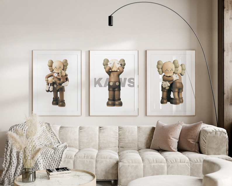 May include: Three framed prints of brown and grey cartoon figures with the word "KAWS" printed in white on the middle print. The prints are hanging on a white wall above a white couch with two pillows.