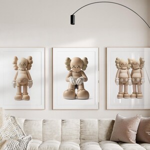 May include: Three framed prints showcasing beige cartoon figures with X-shaped eyes and hands. The figures are in different poses, framed in white, and displayed above a beige sofa with pink cushions. A modern black floor lamp arches over the artwork.