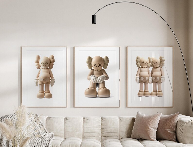 May include: Three framed prints showcasing beige cartoon figures with X-shaped eyes and hands. The figures are in different poses, framed in white, and displayed above a beige sofa with pink cushions. A modern black floor lamp arches over the artwork.