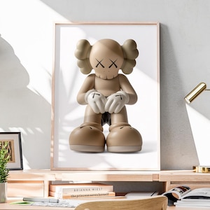 May include: A framed print featuring a tan cartoon figure with X-shaped eyes and a skull-like mouth. The figure has large feet and hands with white gloves. The artwork is displayed on a wooden desk with a small plant and a gold desk lamp.