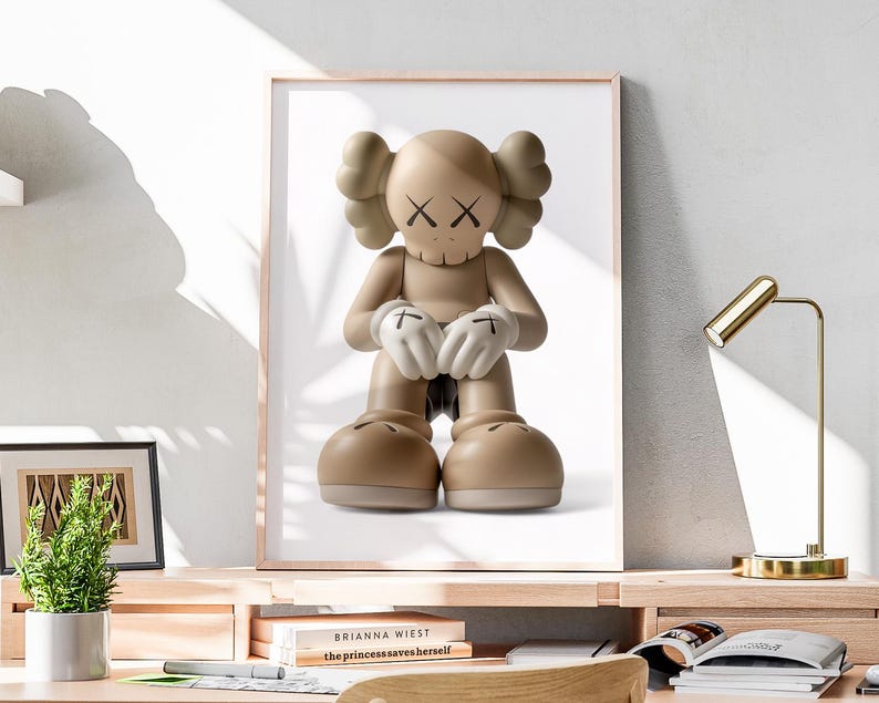 May include: A framed print featuring a tan cartoon figure with X-shaped eyes and a skull-like mouth. The figure has large feet and hands with white gloves. The artwork is displayed on a wooden desk with a small plant and a gold desk lamp.