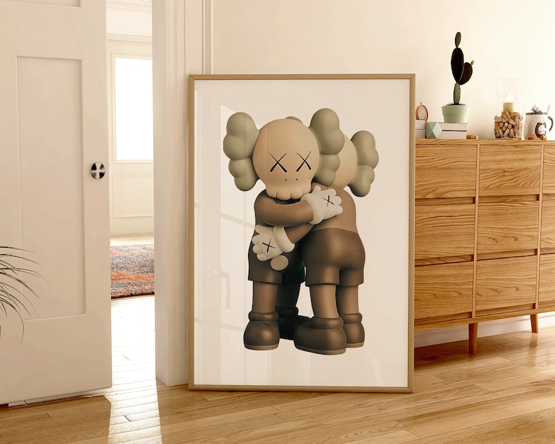 May include: A framed print of two brown and beige cartoon figures hugging each other. The figures have large heads with X-shaped eyes and small mouths. The print is on a white background.