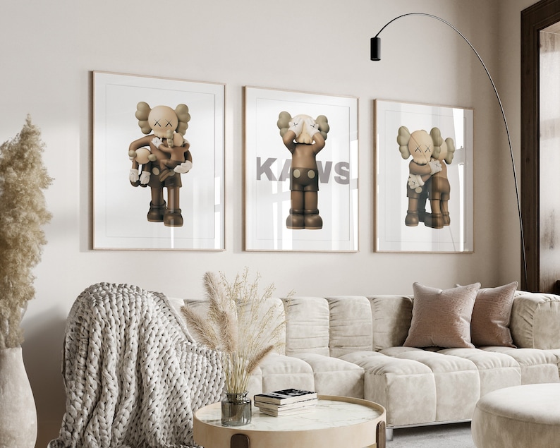 May include: Three framed prints of brown and white cartoon figures with the word "KAWS" printed in black on the middle print.