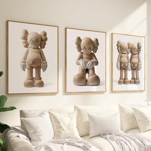 May include: Three framed art prints showcasing a cartoon figure with crossed-out eyes and gloved hands. The figures are in different poses and shades of beige, set against a white backdrop. The frames are gold, and the artwork hangs above a white sofa.