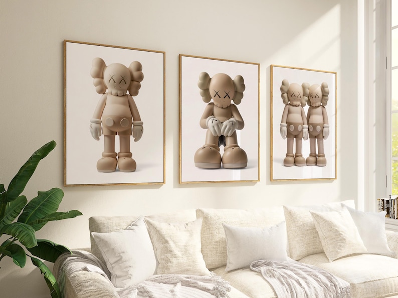 May include: Three framed art prints showcasing a cartoon figure with crossed-out eyes and gloved hands. The figures are in different poses and shades of beige, set against a white backdrop. The frames are gold, and the artwork hangs above a white sofa.